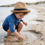 Beach Packing List for Toddlers: What You Actually Need (and What to Skip) Toddler in sun hat and rash guard playing at the beach shoreline