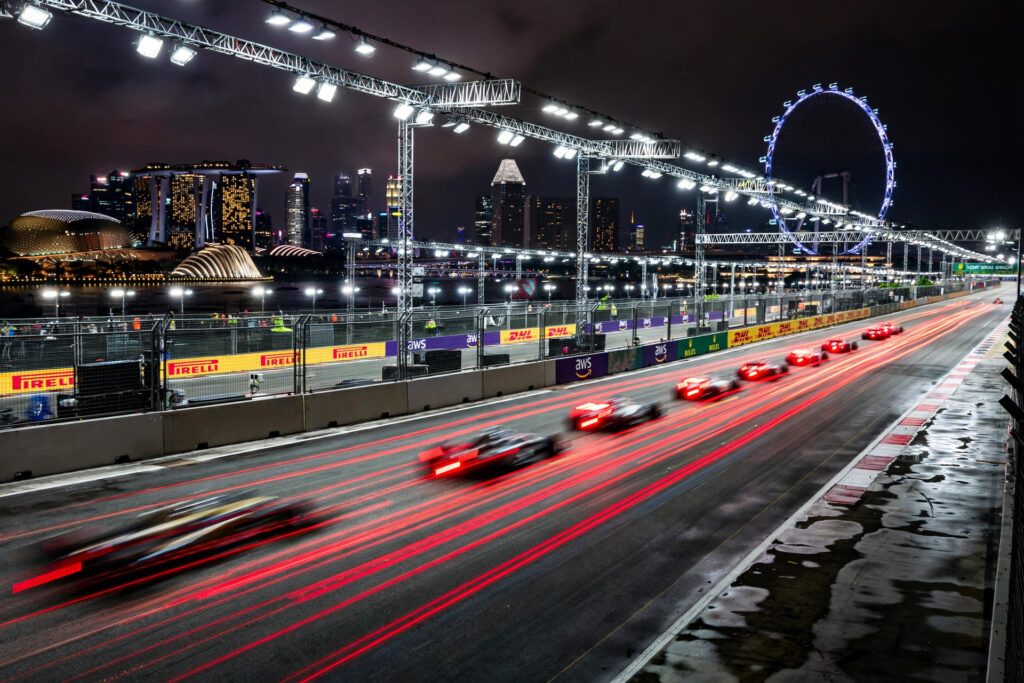 Photorealistic wide landscape of the Marina Bay street circuit at night, Formula 1 race cars with glowing red tail lights streaming under a dense canopy of floodlights, Singapore skyline and the Singapore Flyer behind, rain-free dark sky, no close-up faces