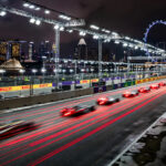 Photorealistic wide landscape of the Marina Bay street circuit at night, Formula 1 race cars with glowing red tail lights streaming under a dense canopy of floodlights, Singapore skyline and the Singapore Flyer behind, rain-free dark sky, no close-up faces
