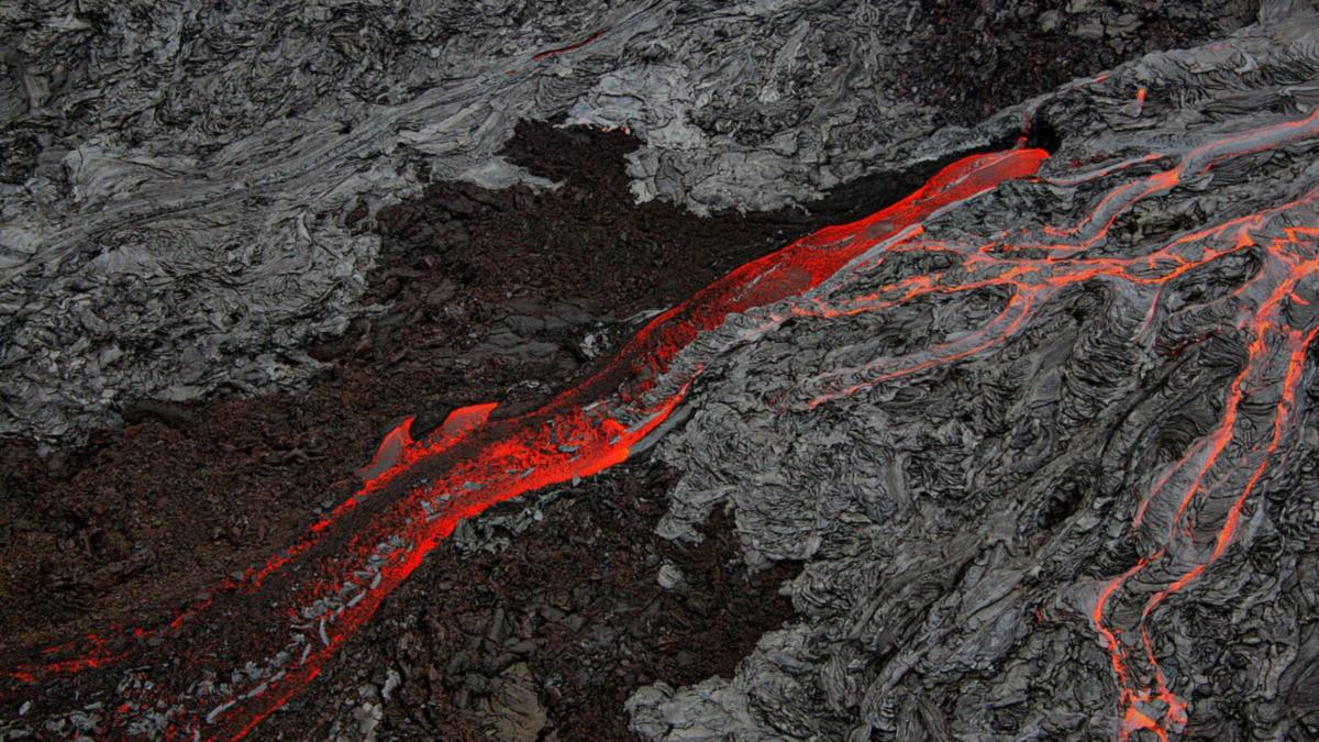 A stream of glowing red lava flowing between dark, cooled lava flows in Hawaii