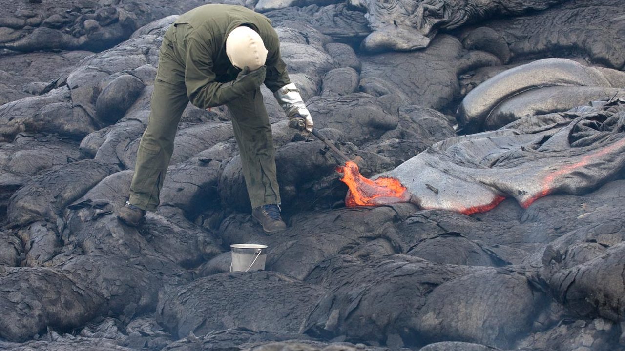 A volcanologist in full protective clothing sampling glowing lava with a hammer and bucket on a cooled lava field