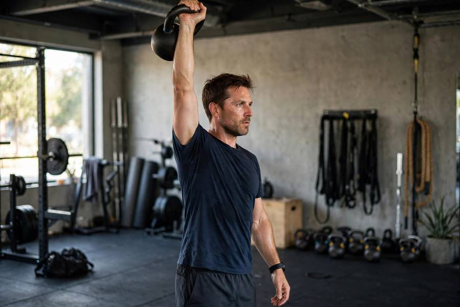 Man performing the standing phase of a Turkish get-up with a kettlebell locked out overhead in a gym