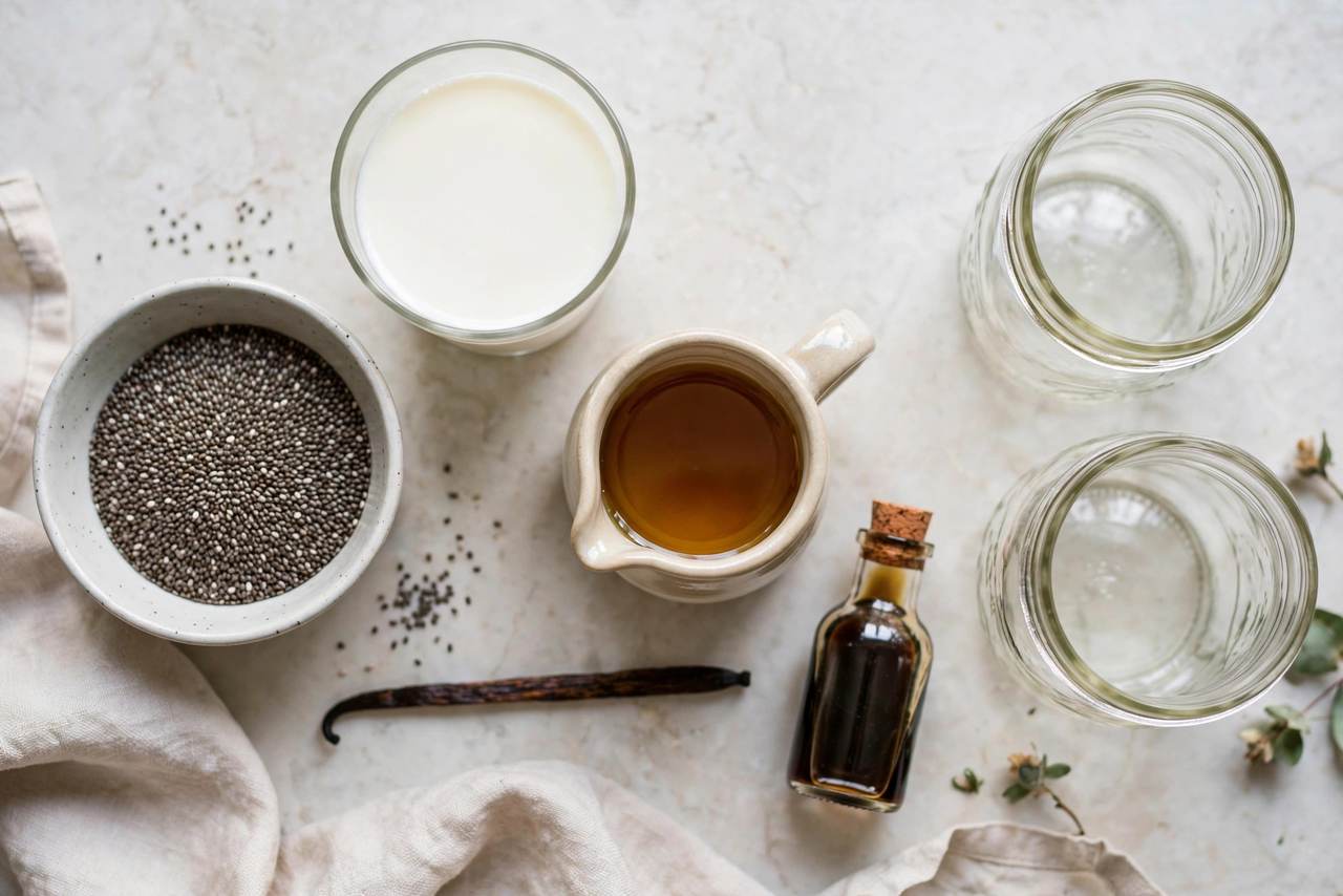 Chia pudding ingredients laid out: chia seeds, milk, maple syrup, vanilla, and mason jars