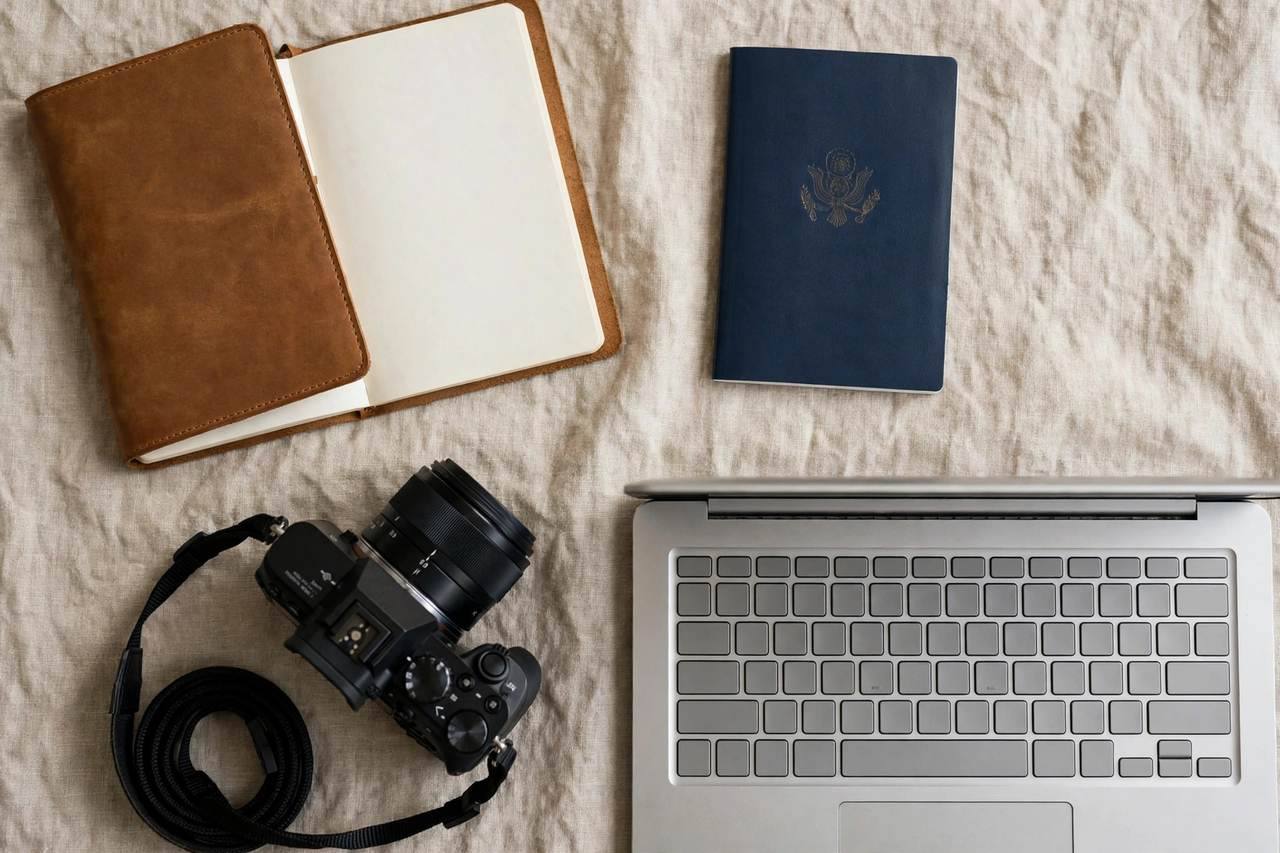 Overhead flat lay of travel planning: notebook, passport, camera, and laptop keyboard