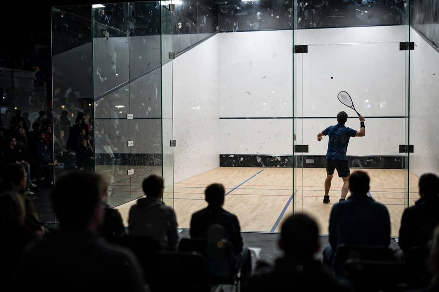 A squash serve seen through the glass back wall of the court