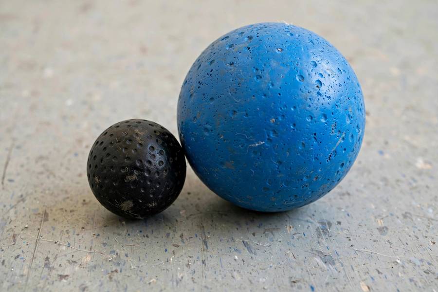 Photorealistic close-up of a small black squash ball next to a larger blue racquetball on a court floor